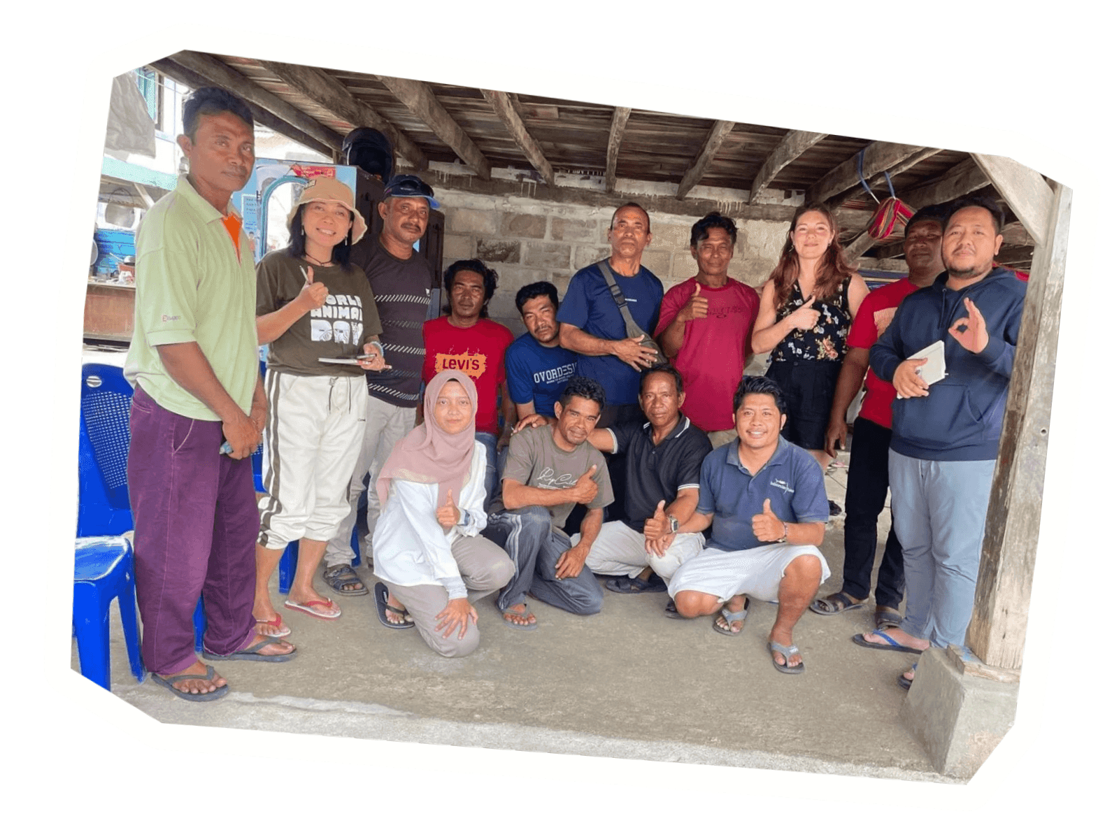 A diverse group of people, including men and women, smiling and gesturing under a rustic shelter.
