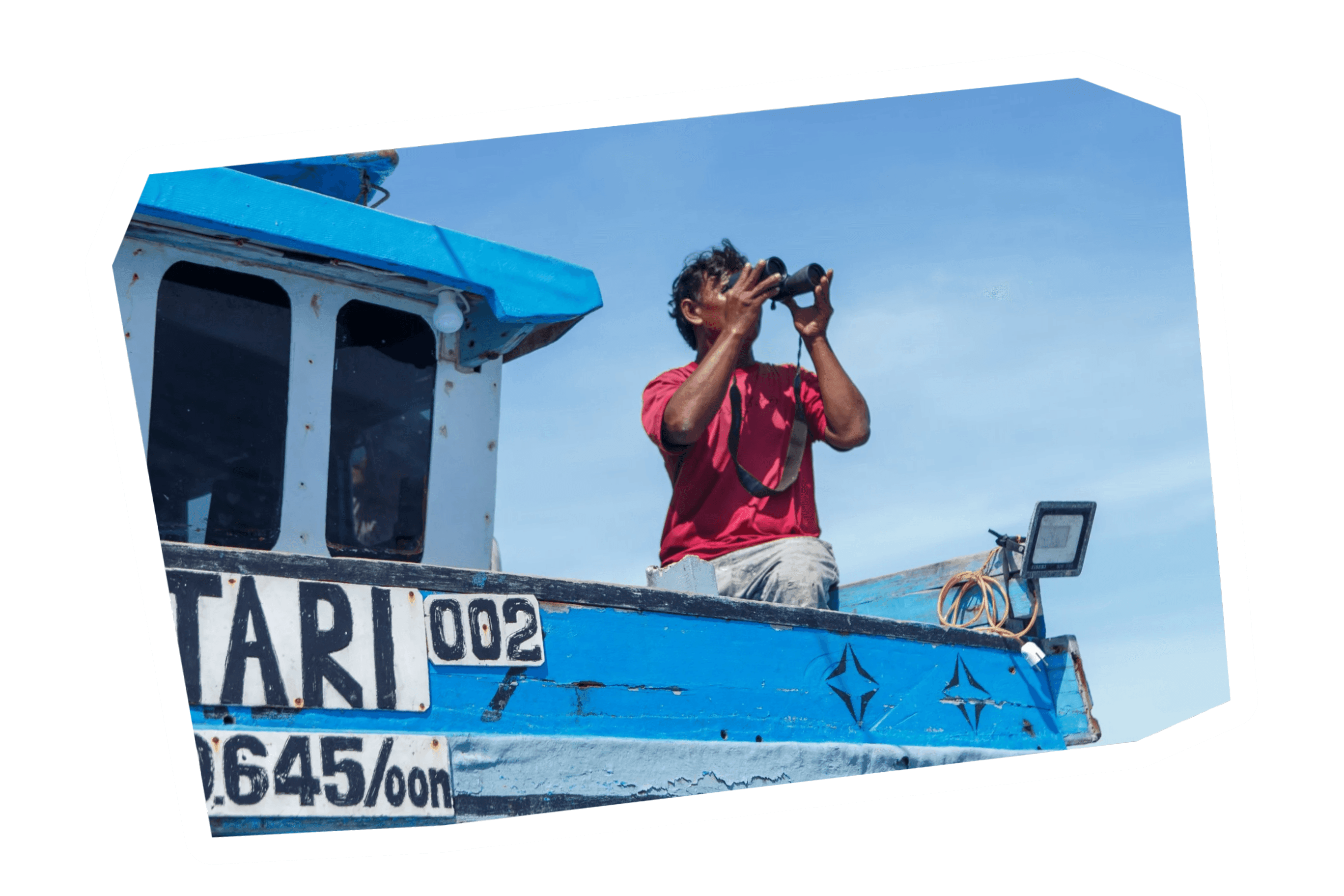 Man in a red shirt on a blue boat looking through binoculars under a clear sky.